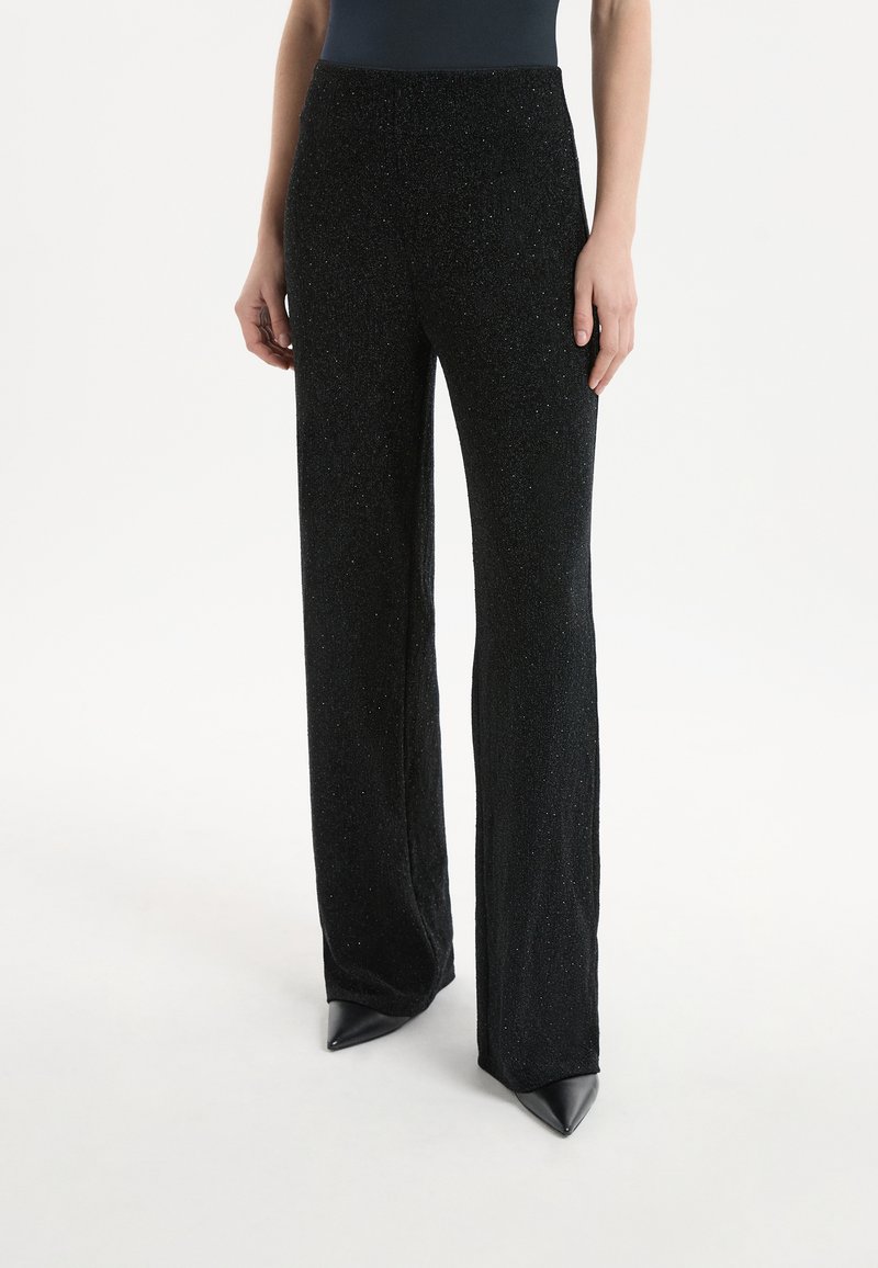 Pantaloni neri scintillanti con design svasato, caratterizzati da una vita alta e una texture morbida. Indossati con scarpe nere a punta.