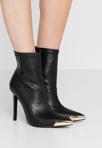 Nogi w czarnych skórzanych botkach na obcasie, z metalowymi złotymi czubkami, stojące na białej powierzchni.