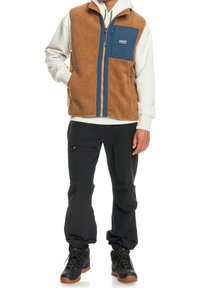 Brun fleeceväst med en blå ficka, som bärs över en grå huvtröja. Svarta byxor och brun-sulade kängor fulländar outfiten.