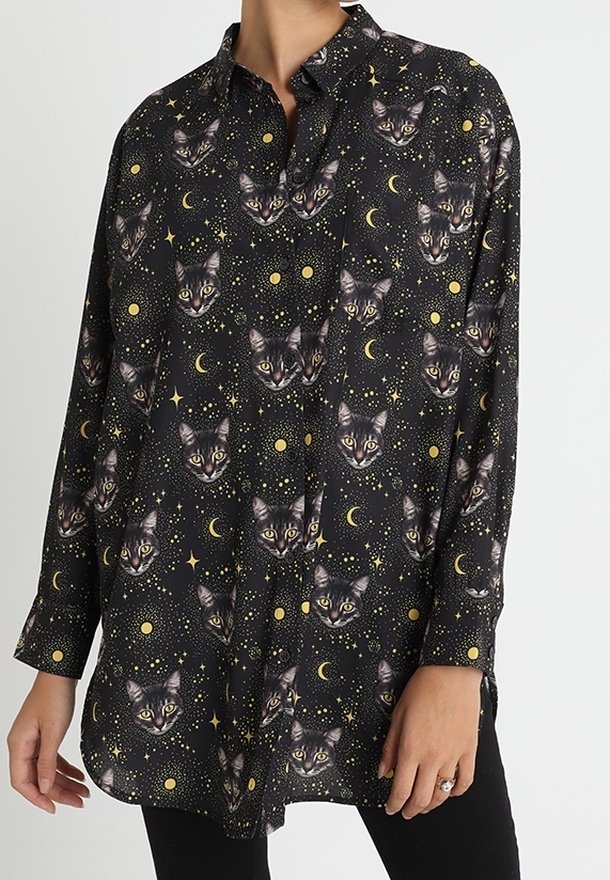 Chemise noire à manches longues boutonnée avec un motif répétitif de visages de chat, lunes jaunes, étoiles et points portée par une personne.