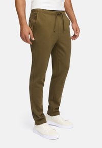 Mann in olivgrünen Jogginghosen mit Kordelzug und weißen Sneakers, der vor einem schlichten Hintergrund steht.