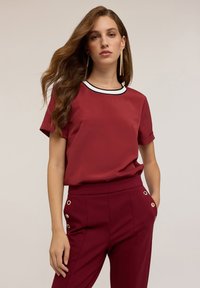 Top bordeaux con maniche corte e collettino bianco e nero, abbinato a pantaloni coordinati con accenti in ottone dorato sui lati.