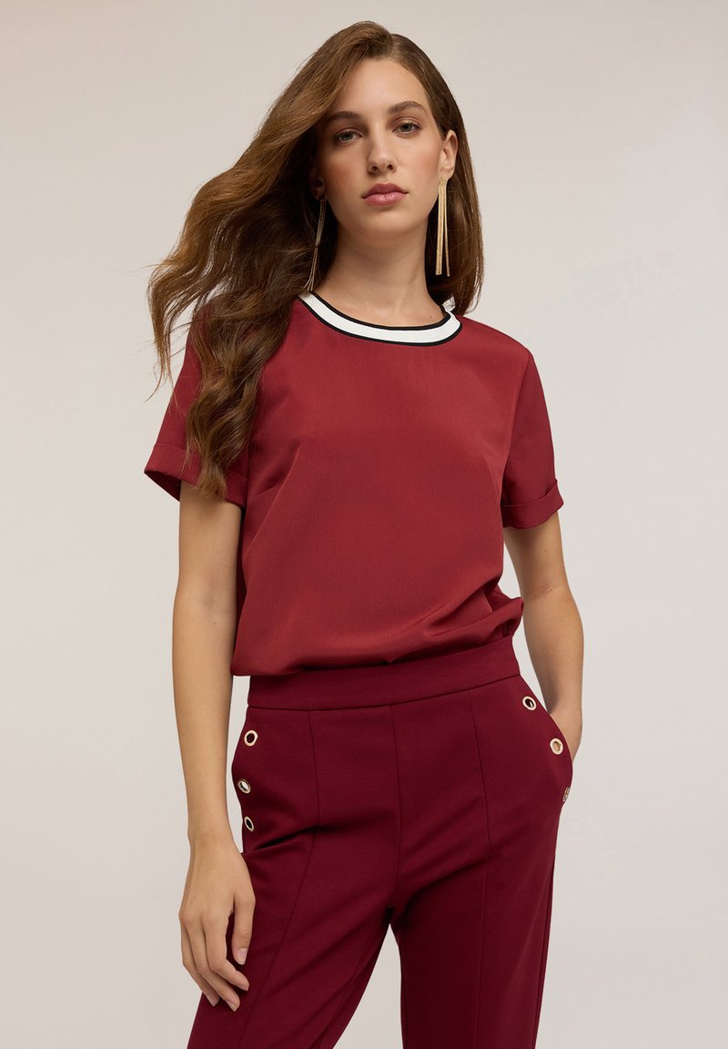 Top bordeaux con maniche corte e collettino bianco e nero, abbinato a pantaloni coordinati con accenti in ottone dorato sui lati.