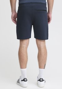 Marineblauwe shorts met een gladde textuur, elastische tailleband en een ritssluiting op de achterzak. Gecombineerd met witte sokken en Adidas sneakers.