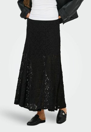 Femme portant une longue jupe noire en dentelle, un haut blanc, une veste en cuir noire et des chaussures noires à enfiler avec des ornements décoratifs.