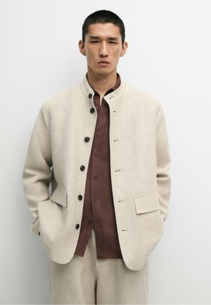 Uomo con capelli corti e scuri che indossa una giacca beige abbottonata sopra una camicia marrone, in piedi davanti a uno sfondo bianco semplice.