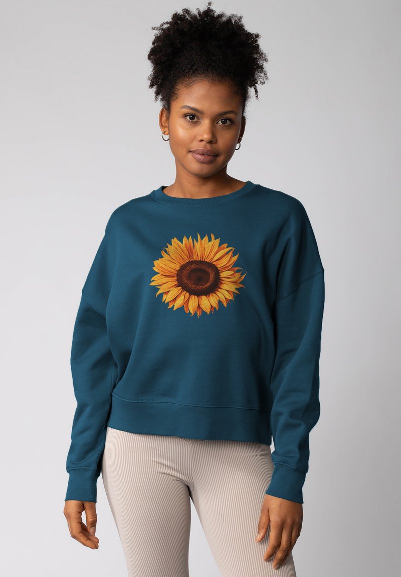 Teal sweatshirt met een levendige zonnebloemafbeelding in het midden. Heeft een relaxte pasvorm, lange mouwen en ribgebreide manchetten. Textuurstof.