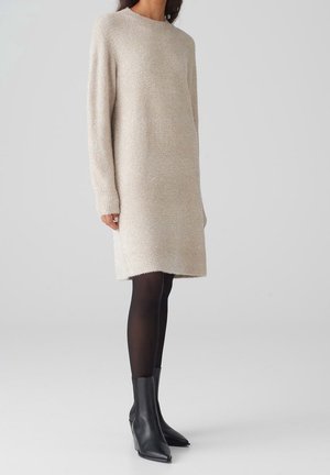 Femme portant une robe-pull tricotée beige à manches longues, des collants noirs et des bottines noires, debout sur un fond clair uni.
