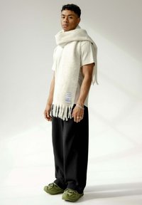 Écharpe en laine crème avec des franges, portée sur un t-shirt blanc. Un pantalon plissé noir et des chaussures vertes complètent la tenue. Design simple et texture douce.