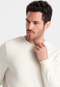 Sweat-shirt de couleur crème avec un col rond et des poignets côtelés, présentant une texture lisse et douce ainsi qu'un design minimaliste.