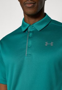 Τιρκουάζ πόλο μπλούζα με υφή, δύο κουμπιά και γκρι γραμμή τόνου κατά μήκος της πατιλέτας. Λογότυπο Under Armour σε γκρι στο στήθος.