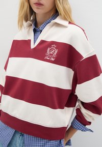 Felpa a righe rosse e crema con scollatura a V e vestibilità oversize. Presenta un logo ricamato e un colletto, indossata sopra una camicia a scacchi blu.