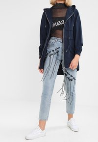 Parka à capuche marine, haut en mesh noir et jeans délavés clairs avec accents graphiques noirs. Des baskets blanches complètent la tenue. Matériaux texturés visibles.