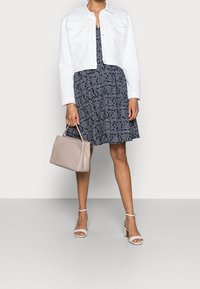 Vit cropped denimjacka över en mörkblå klänning med paisleymönster. Håller en beige handväska och har vita klacksandaler.