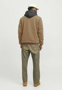 Brun fleecejacka med svart huva, i kombination med urblekta blå jeans och beige kängor. Enkel design, texturerad tyg, och neutral färgskala.