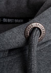 Grauer Hoodie-Stoff mit strukturierter Oberfläche, ausgestattet mit einem Metalldübel und einer dekorativen Lederkordel. Schwarzes Etikett ist innen sichtbar.