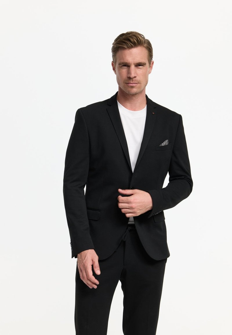 Hombre con chaqueta y pantalones de traje negros sobre una camisa blanca, ajustándose la chaqueta con un fondo blanco neutro.