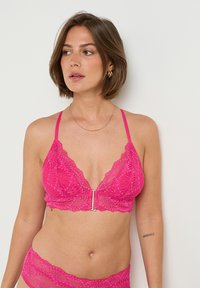 Femme portant de la lingerie en dentelle rose vif avec des boucles d'oreilles en or et un fin collier en or, regardant vers sa gauche contre un fond blanc.
