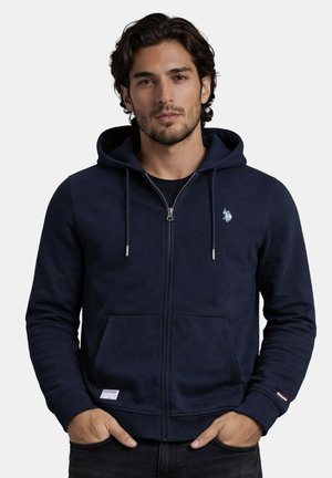 Man with dark hair wearing navy blue zip-up hoodie with hood and front pockets, hands in pockets, facing forward against plain background.