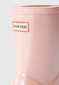 Botte de pluie en caoutchouc rose avec une bordure supérieure texturée, comportant une étiquette rectangulaire blanche avec un contour rouge et un texte noir, "HUNTER".