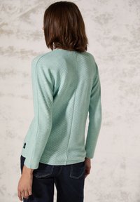 Maglietta in blu-verde chiaro a maniche lunghe con strisce diagonali testurizzate, scollo rotondo e un leggero orlo alto-basso, abbinata a jeans in denim scuro.
