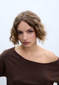 Haut marron en tricot à épaules dénudées avec une texture douce. Le modèle a des cheveux ondulés mi-longs et un maquillage naturel, sur un fond gris clair.