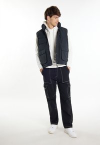 Gilet senza maniche blu navy con multiple tasche frontali, indossato sopra un hoodie bianco, abbinato a pantaloni cargo scuri con cuciture chiare e sneakers bianche.