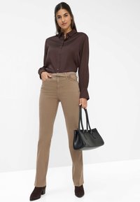 Camicia di seta marrone con bottoni e maniche lunghe, pantaloni slim-fit color sabbia e borsa in pelle nera. Modella con stivaletti alla caviglia marrone scuro.