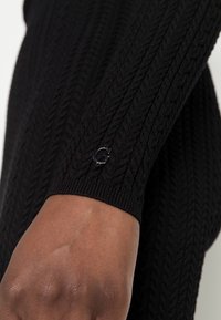 Détail d'un pull en maille noire avec un motif torsadé et des poignets côtelés. Un petit logo argenté est visible sur le poignet.