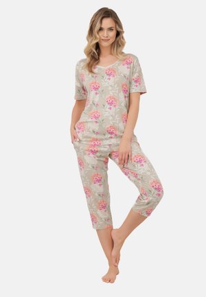 Kobieta stojąca boso, ubrana w beżową piżamę z krótkim rękawem i kwiatowym wzorem oraz dopasowane spodnie 7/8, ręce w kieszeniach, uśmiechnięta.