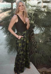 Svart, glittrande bodysuit med djup V-ringning, kombinerad med vida byxor med ett mönster av gröna palmträd. Modellen står mot palmblad.