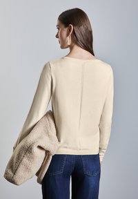 Beige langwerpig top met een ronde hals en een getextureerde achternaad, gecombineerd met een donzige beige buitenjas. Donkerblauwe jeans met zichtbare zakken.