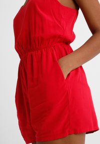 Gros plan d'une personne portant une combinaison rouge sans manches avec une taille élastique et une main dans la poche latérale.