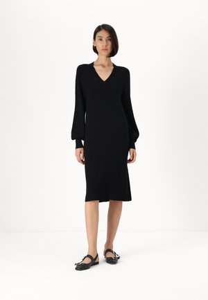 Abito nero lavorato a maglia con maniche lunghe a sbuffo e scollo a V, lunghezza al ginocchio con silhouette dritta. Indossato con scarpe di tela nere con cinturini.