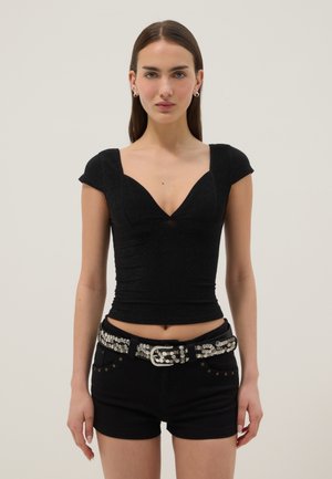 Frau mit glattem braunem Haar trägt ein schwarzes, enganliegendes Top mit Kapuzenärmeln, schwarze Shorts und einen silbernen Nietengürtel und steht vor einem einfarbigen Hintergrund.