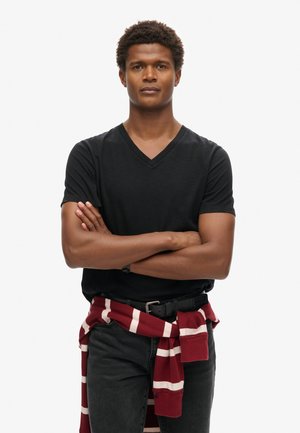 Zwarte V-hals T-shirt met korte mouwen, gecombineerd met donkere spijkerbroek. Een rood en wit gestreepte trui is rond de taille geknoopt.