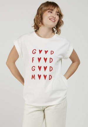 Jeune femme en t-shirt blanc avec des lettres rouges et des formes de cœur disposées en quatre rangées, debout les mains sur les hanches et souriante.