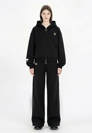 Joven mujer de pie, vistiendo una sudadera negra con capucha y cremallera oversize y pantalones de pierna ancha con tiradores de cremallera circulares y pequeños parches con logo.