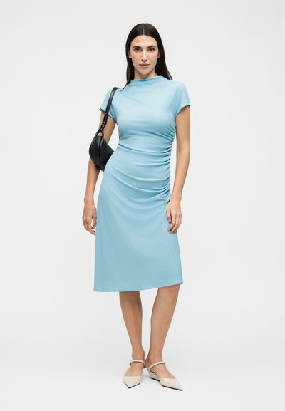 Γυναίκα που φοράει ένα γαλάζιο ρουά μίντι φόρεμα με ριχτά, κρατώντας μαύρη τσάντα ώμου και μπεζ μυτερές μπαλαρίνες, στέκεται μπροστά σε απλό φόντο.