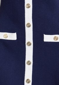 Navy gebreide stof kledingstuk met witte accenten, acht metalen knopen in het midden en twee witte zakflappen.