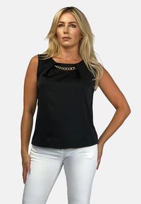 Czarny bezrękawnik z plisą i złotym łańcuszkiem przy dekolcie; gładki materiał i luźny krój, w połączeniu z białymi spodniami.