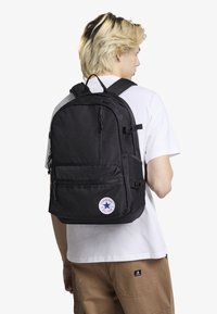 Mochila de tela negra con múltiples cremalleras, correas ajustables y un parche de logo circular. Presenta una superficie texturizada y un diseño estructurado.