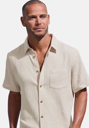 Light beige short-sleeve shirt with a textured fabric, a front pocket, and bronze buttons. Collared design with a relaxed fit.