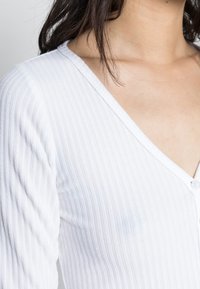 Žena v bílé, žebrované, dlouhé rukávy s výstřihem do V a detailem knoflíku, s tmavými vlasy částečně viditelnými přes rameno.