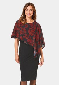 Robe noire avec une superposition transparente présentant des motifs floraux rouges et des bordures noires, créant un look empilé. Le mannequin se tient debout, montrant la tenue.