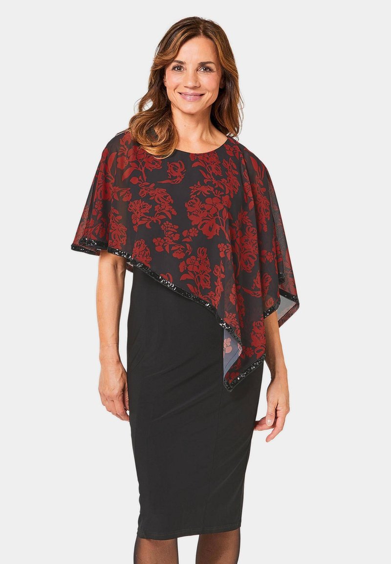 Robe noire avec une superposition transparente présentant des motifs floraux rouges et des bordures noires, créant un look empilé. Le mannequin se tient debout, montrant la tenue.