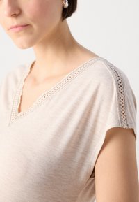 Beige kortef Mouwen top met een V-hals en kantafwerking langs de schouders, gemaakt van een zachte, lichte stof met een gladde textuur.