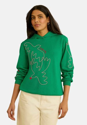 Kobieta w zielonej bluzie z kapturem z czerwono-białym rysunkiem ptaka na przodzie i rękawie, w połączeniu z beżowymi spodniami, stojąca na białym tle.