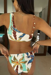 Több színű mintával díszített bikini, amely pálmaleveleket ábrázol. Egy vállas felsőrész, díszes kagyló részletekkel és vállpántokkal. Pamut keverékből készült anyag.