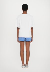 Hvit kortermet t-skjorte, blå stripete shorts og sølvfargede sko. Bakgrunnen er ensfarget hvit, noe som fokuserer på fargene og designet til antrekket.